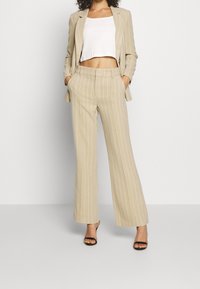 Beige, gestreifter Anzug mit taillierter Blazerjacke und weit geschnittenen Hosen, kombiniert mit einem weißen Crop-Top und schwarzen High-Heels.