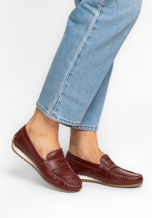 Person trägt hellblaue Jeans und dunkelrote Leder-Loafer mit weißen Sohlen, steht vor einem schlichten weißen Hintergrund.