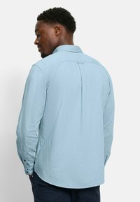 Himmelblaues Langarmhemd aus strukturiertem Stoff, mit geknöpften Manschetten und einer Rückenfalte. Kragen- und abgerundeter Saum-Detail.