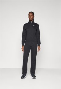 Veste en polaire avec fermeture éclair noire accompagnée d'un pantalon assorti. Texture lisse, design ajusté, minimalisme de la marque sur la poitrine, et baskets à semelle en caoutchouc.