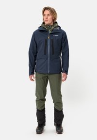 Giacca impermeabile blu navy con dettagli neri, due tasche sul petto e cerniera frontale. Abbinata a pantaloni verde oliva e gaiters neri.