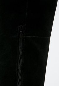 Bota de ante negra por encima de la rodilla con cierre de cremallera en el lateral, textura suave y un diseño ajustado que se adapta a la pierna.