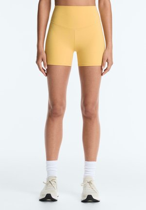 Nő mustársárga, magas derekú sportnadrágot, fehér középmagasságú zoknit és bézs futócipőt visel, karjai laza tartásban áll.