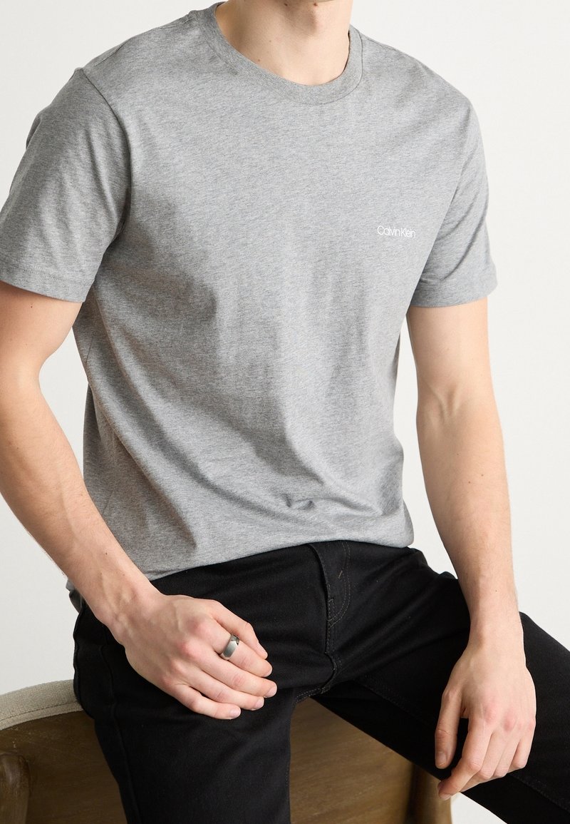 T-shirt gris en coton avec un col rond et des manches courtes, arborant un petit logo blanc Calvin Klein sur la poitrine. Un jean noir et une bague sont visibles.