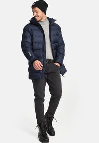 Chaqueta de plumas azul marino con cremallera frontal, capucha y puños elásticos, llevada sobre una camiseta gris, combinada con jeans negros y botas de cordones negras.