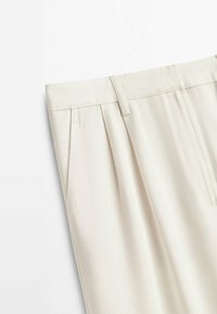 Gros plan sur un pantalon beige clair montrant la ceinture, les passants et les plis latéraux sur un fond blanc uni.