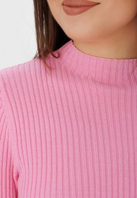 Haut côtelé ajusté rose, avec un col montant et un tissu textur é. Un détail en gros plan met en évidence la douceur du matériau.