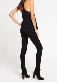 Svart ärmlös topp med lös passform, kombinerad med tajta svarta jeans och svarta klackboots i ankellängd, sedd bakifrån.