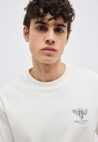 Νεαρός με σκούρα σγουρά μαλλιά που φοράει λευκό φούτερ με λογότυπο μέλισσας και την επιγραφή "Marc O'Polo" στο στήθος.