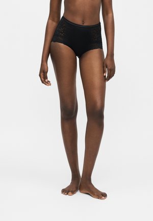 Kvinde model iført sort højtaljet undertøj med blonder på siderne, stående barfodet mod en ensartet lysegrå baggrund.