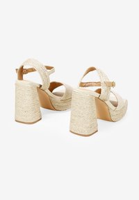 Sandalias de plataforma beige con correas anchas y trenzadas y tacones cuadrados con textura de paja. Presenta una correa ajustable en el tobillo para un ajuste seguro.