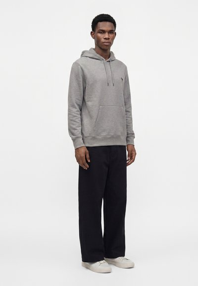Homme debout portant un sweat à capuche gris, un pantalon noir et des baskets blanches sur un fond gris clair uni.