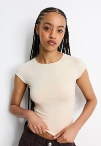 Beige eng anliegendes T-Shirt mit kurzen Ärmeln. Hergestellt aus glattem, leichtem Stoff. Minimalistisches Design mit rundem Ausschnitt und ohne sichtbare Muster.
