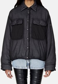 Czarna, ocieplana kurtka koszulowa z dużymi kieszeniami na piersi, zapinana na guziki, z elastycznym dolnym brzegiem, noszona na warstwę na koszulę dżinsową. Tkanina o fakturze.