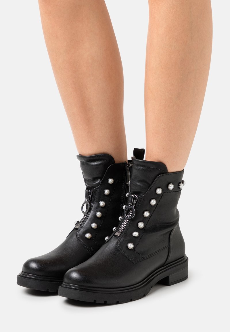 Bottines en cuir noir avec fermeture éclair sur le devant et clous décoratifs argentés portées sur jambes nues sur fond blanc.