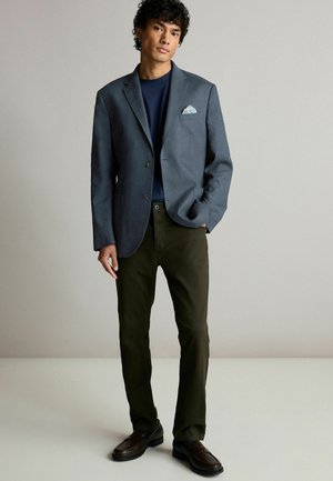 Blazer gris à deux boutons et poches latérales, associé à une chemise marine et un pantalon vert foncé. Des chaussures marron complètent le look.