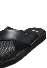Czarna skórzana sandałka na płaskiej podeszwie z plecionym teksturowanym paskiem oraz wytłoczonym logo BOSS na górnej części i wkładce.