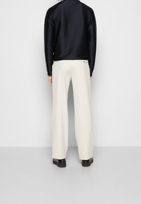 Person som bär en svart långärmad topp, lösa vita byxor med knäppflik på bakfickan och svarta skor, står mot en ensam bakgrund.