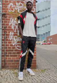 Tepláková souprava má černou bundu s červenými akcenty a bílými pruhy; černé kalhoty s červenými a bílými diagonálními vzory; bílé tenisky.