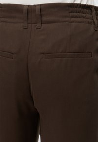 Gros plan de la ceinture d'un pantalon marron avec passants de ceinture et deux poches arrière passepoilées sur un tissu à texture sergée diagonale.