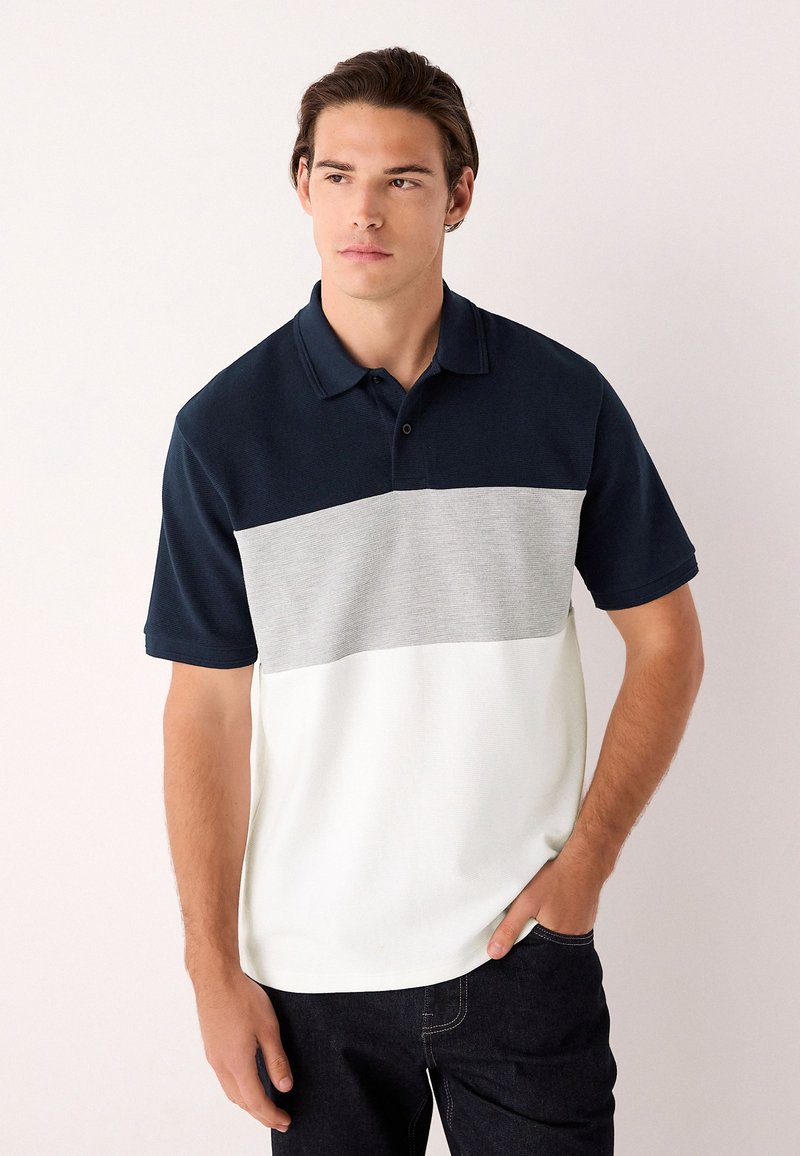 Poloshirt in Marineblau, Grau und Weiß. Kurze Ärmel, Kragen und ein Color-Block-Design mit strukturierter Stoffqualität. Kombiniert mit dunklen Jeans.