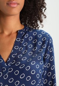 Blouse bleu marine avec un col en V et un motif circulaire blanc. Texture de tissu douce, avec une fermeture à boutons au niveau du col.