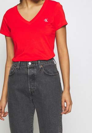 Kvinde iført en kraftigt rød T-shirt med V-hals med et lille hvidt "cK"-logo og højttaljede sorte vaskede jeans.