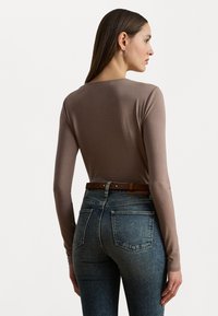 Langarmshirt aus taupefarbenem Baumwollgemisch, kombiniert mit hochgeschnittenen blauen Jeans. Auffällige Details sind ein brauner Gürtel und eine figurbetonte Silhouette.
