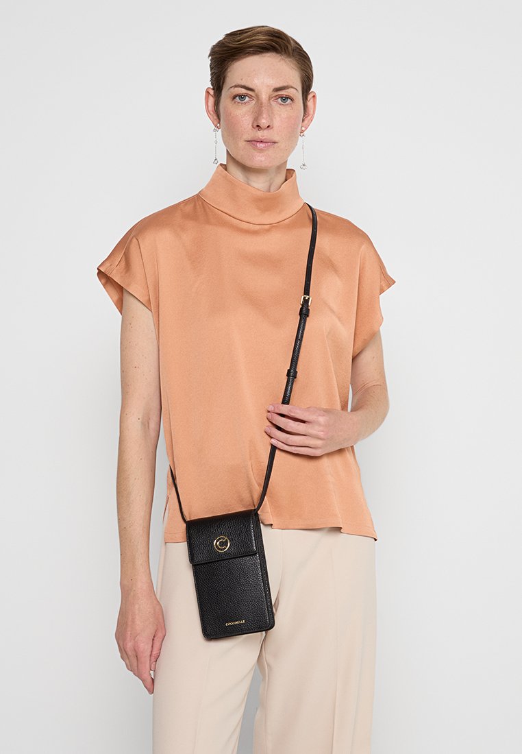 Schwarze, strukturierte Leder-Umhängetasche mit goldenen Hardware-Details, getragen über einem pfirsichfarbenen Satin-Oberteil. Schlichtes Design und rechteckige Form.