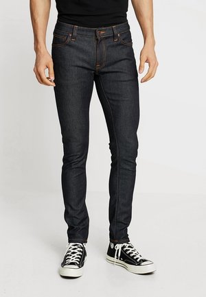 Mies, jolla on tummat kapeat farkut ja mustat korkeavartiset lenkkarit, joissa on valkoiset nauhat, seisoo vaaleaa yksinkertaista taustaa vasten.