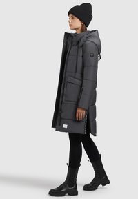 Woman wearing a long dark gray padded coat with hood, black knit beanie, black leggings, and black chunky boots, standing in profile.