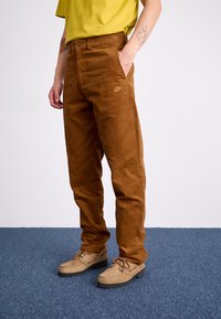 Pantaloni din catifea înnuțită maro, cu croială dreaptă, un logo Nike subtil pe partea stângă și buzunare laterale, asortați cu pantofi bej deschis.