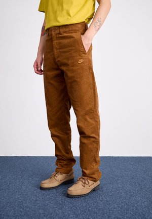 Brown corduroy pants with a straight fit, a subtle Nike logo on the left side, and side pockets, paired with light beige shoes.