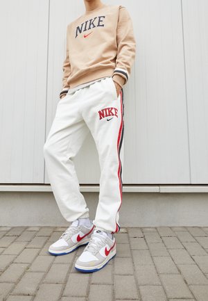 Osoba ubrana w beżową bluzę Nike i białe spodnie dresowe Nike z czerwono-czarnymi paskami, stojąca na wybrukowanym podłożu w adidasach.