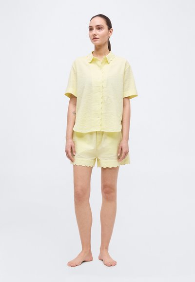 Γυναίκα όρθια, ξυπόλυτη, φοράει ανοιχτό κίτρινο υφασματιστό πουκάμισο με κοντά μανίκια και κουμπιά, και σορτς με ίδια απόχρωση και κυματιστό τελείωμα, σε λευκό φόντο.