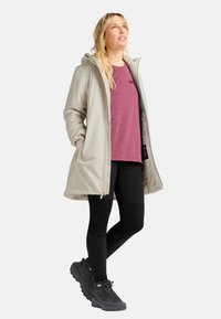 Donna con un cappotto beige con cappuccio, una maglietta viola, leggings neri e scarpe da trekking nere, in piedi e che guarda verso l'alto su uno sfondo bianco.
