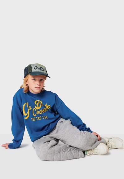 Felpa blu con testo giallo, pantaloni della tuta grigi trapuntati e cappellino verde con dettagli. Sneaker bianche completano l'outfit casual.
