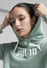 Zelena dukserica s kapuljačom od mekanog materijala, na prednjoj strani ima veliki bijeli "PUMA" logo i ikonu, s vezicom i rebrastim rubom.