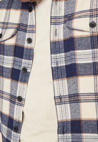 Camicia in flanella con un motivo a quadri blu navy, crema e arancione, caratterizzata da bottoni neri, due tasche sul petto e un tessuto morbido e texture. 