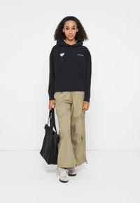 Zwarte hoodie met hartafbeelding en logo, beige cargopants, draagt een zwarte totebag, draagt grijze sneakers, krullend haar.
