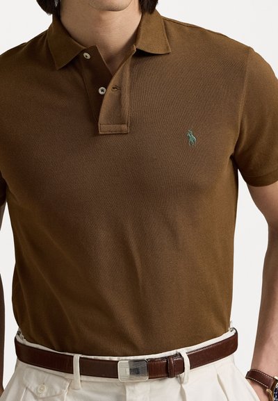 Brūns polo krekls, kas izgatavots no teksturēta auduma, ar divu pogu aizdari un mazu zaļu izšūtu logotipu uz krūtīm. Redzams brūns jostas siksna.