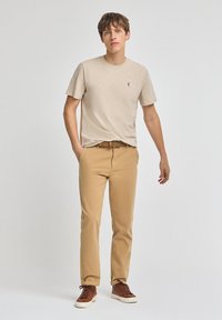 T-shirt in cotone beige con collo rotondo e piccolo logo, abbinata a pantaloni in cotone beige e sneakers marroni con lacci. Design semplice e casual.