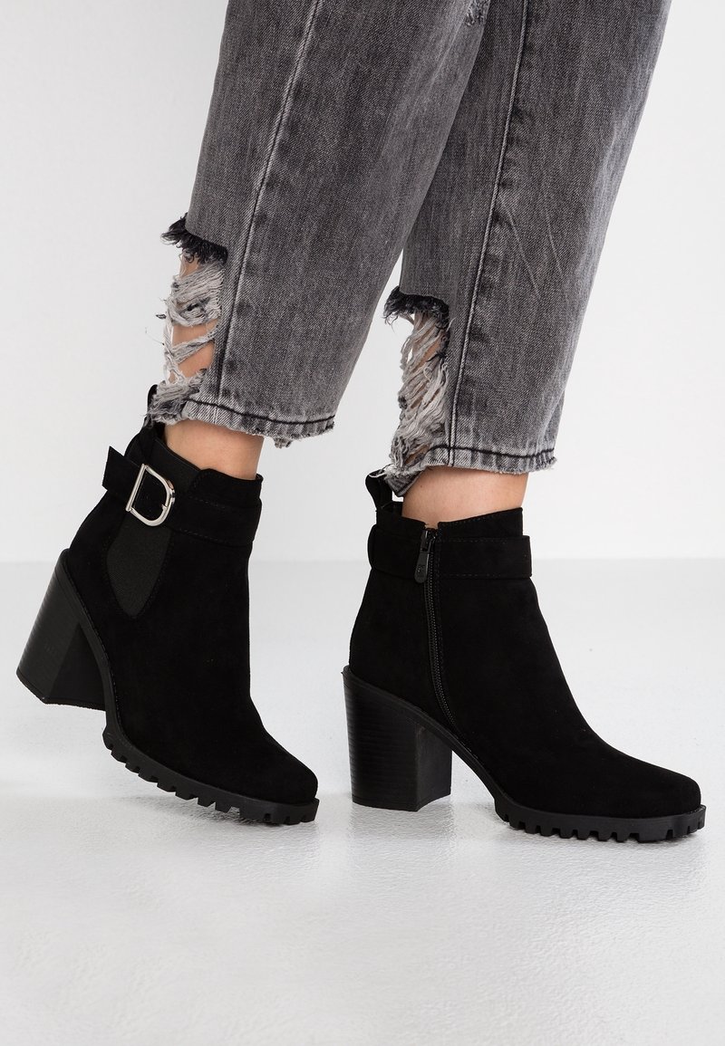 Bottes en daim noir longueur cheville avec talons épais et boucles latérales portées avec un jean gris délavé et court.