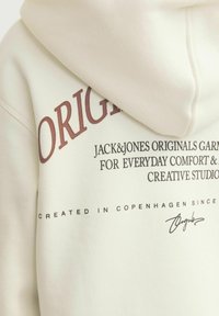 Cream hoodie with a large "ORIGINAL" print in burgundy, and black text detailing. Features a soft texture and kangaroo pocket at the front.