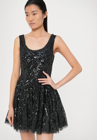 Mladá žena v bezramenných černých mini šatech s flitrovou výšivkou, ruka na bocích, na jednoduchém světlém pozadí.