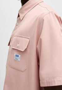 Chemise rose clair à manches courtes avec col, dotée d'une poche poitrine et d'une étiquette de marque. Tissu texturé avec fermeture à boutons.