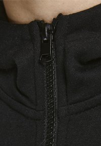 Nära bild av en svart dragkedja på ett mörkt tygplagg, delvis draget vid halslinningen på en person.