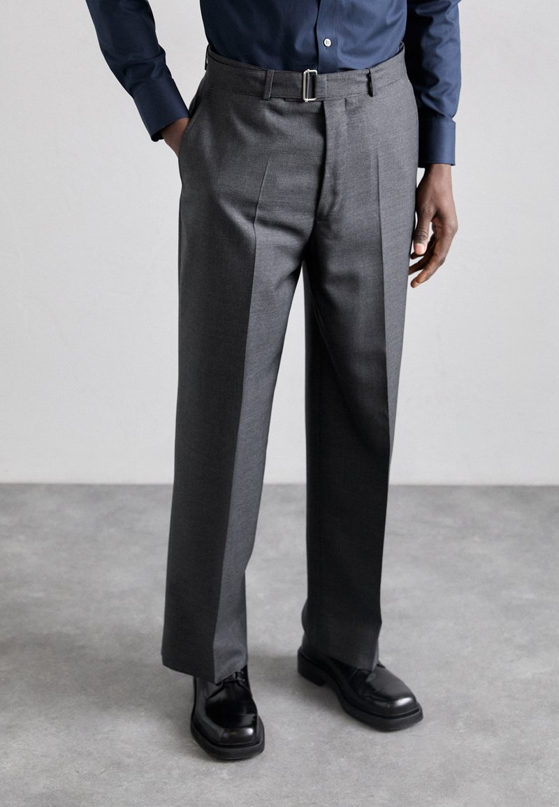Pantalons gris ajustés en tissu mélangé de laine, avec une taille plate, des passants de ceinture et des plis subtils. Associés à des chaussures en cuir noires.