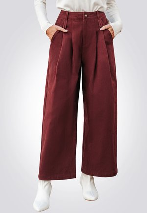 Brede pijpenbroek van bordeauxrood stof, met voorplooien, zijzakken en een enkele knoopsluiting, gecombineerd met witte enkellaarsjes.
