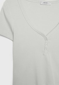 Ljusgrå kortärmad t-shirt med djup V-ringning, med fyra vita knappar fram och en slät, mjuk textur.